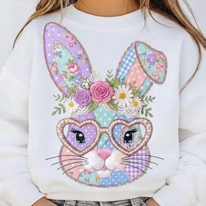 Peut inclure: Sweat-shirt blanc avec un motif de visage de lapin patchwork. Le lapin porte des lunettes en forme de cœur et des détails floraux sur les oreilles et une couronne de fleurs. Le design utilise des couleurs pastel et des détails complexes, parfait pour Pâques ou le printemps.