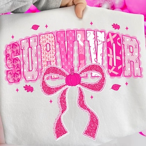May include: White sweatshirt with the word "SURVIVOR" in pink, decorated with pink ribbons and a large pink bow. The sweatshirt is held up by a person.