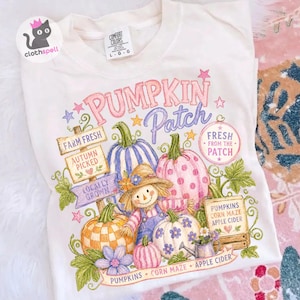 May include: A cream-colored t-shirt featuring a colorful graphic print with the text PUMPKIN Patch, FARM FRESH, AUTUMN PICKED, LOCALLY GROWN, FRESH FROM THE PATCH, PUMPKINS, CORN MAZE, and APPLE CIDER, depicting pumpkins, a scarecrow, and stars.