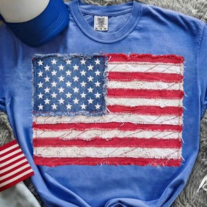 Op de afbeelding: Een lichtblauw T-shirt met een versleten Amerikaans vlagontwerp. De vlag is gemaakt van denim en rood en wit stof, met een gerafelde rand. Een blauw-witte baseballpet is zichtbaar op de achtergrond.