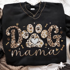 May include: Black sweatshirt with the words "DOG MAMA" in a sparkly, leopard print design. The paw print in the center has a heart. Gold necklace is draped over the sweatshirt.