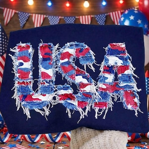 Può includere: Maglietta blu navy con la scritta "USA" in un design patchwork invecchiato. Le lettere sono realizzate in tessuto rosso, bianco e blu, con bordi sfilacciati. Adatta per un tema patriottico.