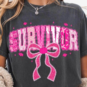 May include: Dark gray t-shirt with the word "SURVIVOR" in pink letters, decorated with pink ribbons and a large pink bow. The shirt is a casual style, suitable for everyday wear.