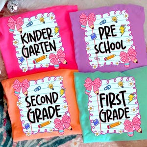 May include: Four colorful t-shirts with school-themed designs. Each shirt features a different grade level: Kindergarten, Preschool, Second Grade, and First Grade. The designs include pencils, bows, and the grade level text.