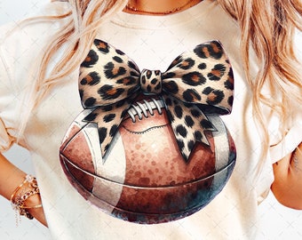 May include: A white t-shirt with a brown football decorated with a leopard print bow. The football is centered on the shirt.