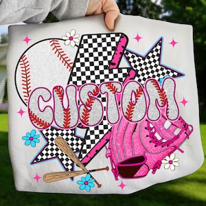 Puede incluir: Bolso tote blanco con un diseño de béisbol, guante y bates cruzados. La palabra "CUSTOM" está en letras brillantes. El diseño incluye elementos rosas, negros y blancos, con estrellas y flores.