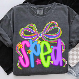 以下が含まれることがあります： カラフルな文字で「SPED」と書かれたグレーのTシャツ。虹色のリボンと星で飾られています。黒いジャケット、デニムショーツ、携帯電話ケースと一緒に表示されています。