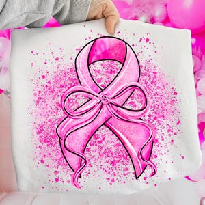 May include: A white tote bag featuring a large pink ribbon design, a symbol of breast cancer awareness. The ribbon is detailed with a leopard print pattern and outlined in black. Pink paint splatters surround the ribbon.