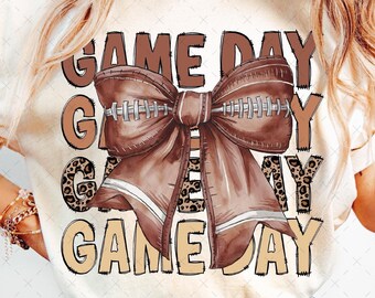 May include: A white t-shirt with a brown and white football-themed design. The design features a large brown bow with a football pattern and the text "GAME DAY GAME DAY".