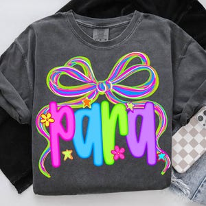 May include: Dark gray t-shirt with a colorful bow and the word "PARA" in bright, playful letters. The design includes stars and flowers. The shirt is paired with denim shorts and a phone case.