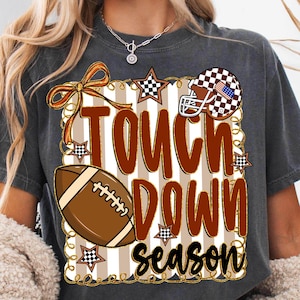 May include: Dark gray t-shirt with a football-themed design. The design includes a football, a helmet, stars, and the words "Touch Down Season" in brown and black. The background has vertical stripes.