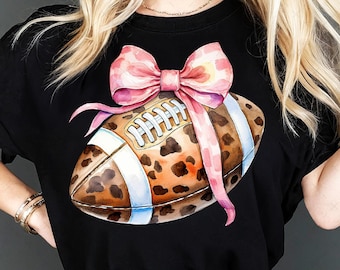 May include: Black t-shirt featuring a watercolor illustration of a football with a leopard print design, white stripes, and a pink bow. The text "PNG DIGITAL DOWNLOAD" is at the bottom.