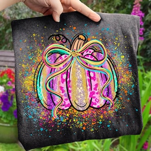 May include: A dark gray t-shirt featuring a colorful pumpkin design. The pumpkin has a pink leopard print pattern, gold glitter accents, and a large bow. The design includes a splatter of colorful paint.