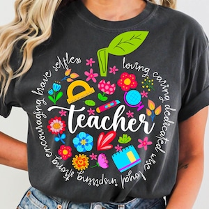 May include: Dark gray t-shirt with a colorful apple design. The apple is composed of flowers, school supplies, and the word "Teacher" in white script. The design includes words like "brave", "loving", and "inspiring".