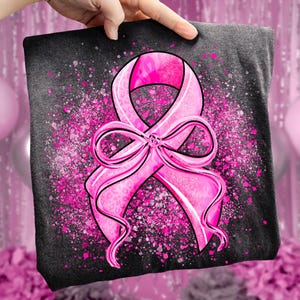 May include: A dark gray t-shirt featuring a large pink ribbon design, a symbol of breast cancer awareness. The ribbon is detailed with a leopard print pattern and a bow. Pink speckles surround the ribbon, creating a textured effect.