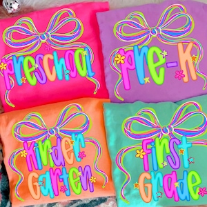 May include: Four colorful t-shirts with school grade level designs. The shirts are pink, purple, orange, and teal. Each shirt has a bow design and the words "Preschool", "Pre-K", "Kindergarten", and "First Grade" in bright colors.