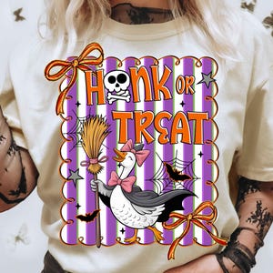 May include: Cream-colored t-shirt with a Halloween-themed graphic. The graphic features a goose wearing a cape and holding a broom, with the words "Honk or Treat" in orange and white. The design includes bats, spiderwebs, and a skull.