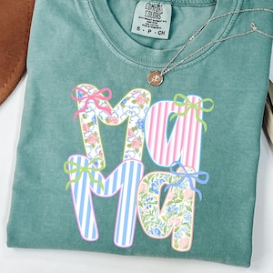 May include: A teal Comfort Colors t-shirt with the word "Mama" in a floral and striped design. The letters are decorated with bows in pink, blue, and green. A gold necklace with a small pendant is draped across the shirt.