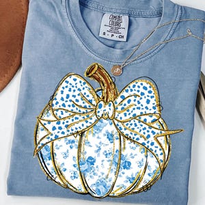 May include: A blue short-sleeved t-shirt featuring a graphic of a pumpkin with a blue and white floral pattern, a blue and white leopard print bow, and gold glitter outlines. The shirt has a Comfort Colors brand label showing size S-CH.