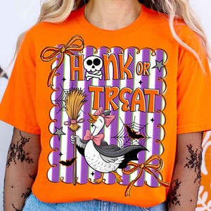 May include: Orange t-shirt with a Halloween-themed graphic. The design features the text "Honk or Treat" with a skull and crossbones, a cartoon goose in a witch costume, bats, and a broom. The graphic is set against a purple and white striped background.