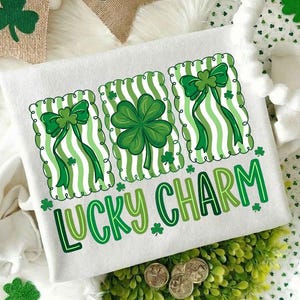 May include: White sweatshirt with a green and white striped design featuring shamrocks and bows. The text "LUCKY CHARM" is printed in green. Decorative shamrock banners and scattered shamrock confetti are also visible.