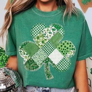 May include: Green t-shirt featuring a patchwork shamrock design. The shamrock is composed of various green and white patterned fabrics, including gingham, floral, and paisley prints. The shirt is a solid green color.