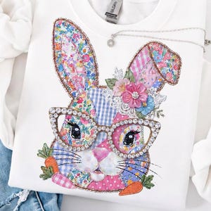 May include: White crewneck sweatshirt featuring a colorful patchwork bunny design. The bunny has large, embellished glasses, floral accents, and carrot details. A silver necklace with a small pendant is draped on the sweatshirt.