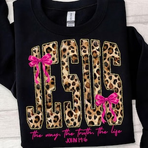 May include: Black sweatshirt featuring the word "JESUS" in a leopard print design, edged with gold. Pink bows are placed on the letters. The text "the way, the truth, the life" and "JOHN 14:6" are in pink.