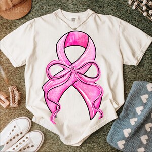 May include: Cream-colored t-shirt featuring a large pink ribbon design with a black outline. The ribbon has a leopard print pattern and is tied in a bow. The shirt is on a green surface with white sneakers and a blue blanket.
