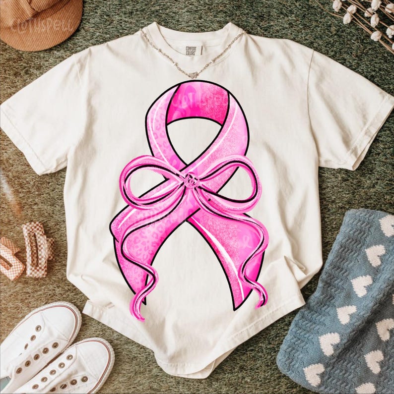 May include: Cream-colored t-shirt featuring a large pink ribbon design with a black outline. The ribbon has a leopard print pattern and is tied in a bow. The shirt is on a green surface with white sneakers and a blue blanket.