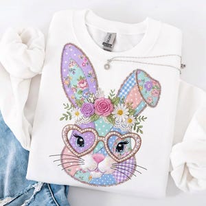 Puede incluir: Sudadera blanca con un dise&ntilde;o de conejo de patchwork colorido con gafas en forma de coraz&oacute;n. El conejo tiene detalles florales y una nariz rosa. Un collar plateado con un peque&ntilde;o colgante descansa sobre la sudadera. Dise&ntilde;o para Pascua.