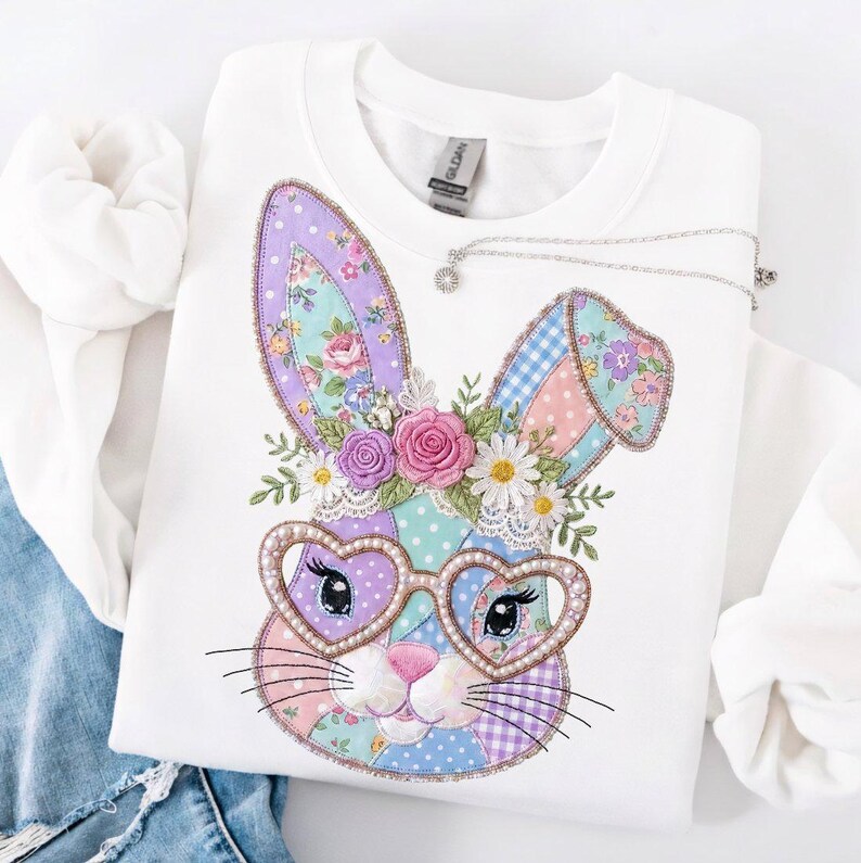 Puede incluir: Sudadera blanca con un dise&ntilde;o de conejo de patchwork colorido con gafas en forma de coraz&oacute;n. El conejo tiene detalles florales y una nariz rosa. Un collar plateado con un peque&ntilde;o colgante descansa sobre la sudadera. Dise&ntilde;o para Pascua.