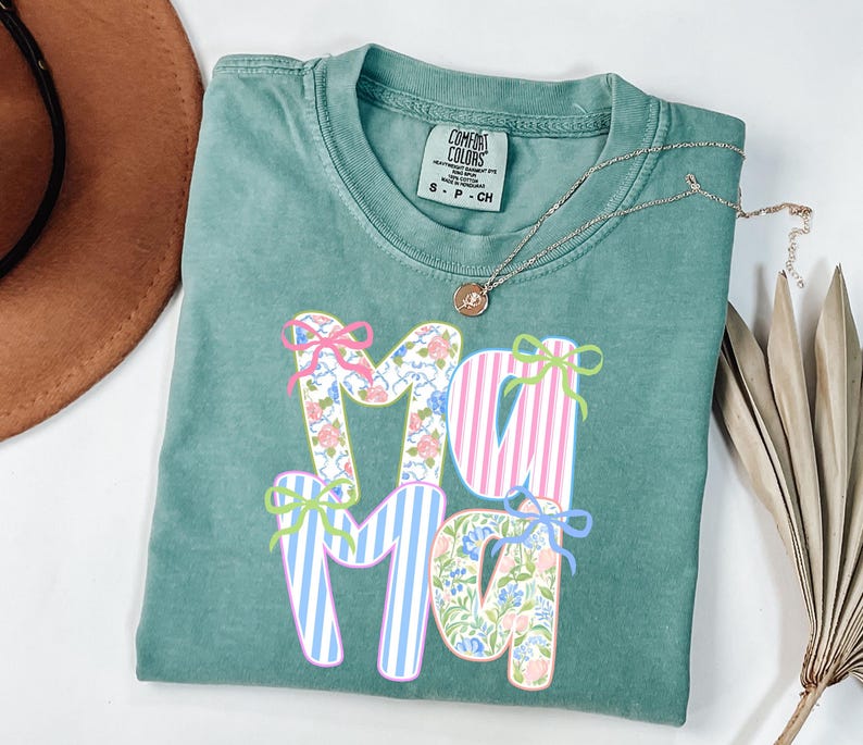 May include: A teal Comfort Colors t-shirt with the word "Mama" in a floral and striped design. The letters are decorated with bows in pink, blue, and green. A gold necklace with a small pendant is draped across the shirt.