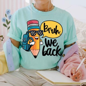 May include: A light teal t-shirt featuring a cartoon pencil character wearing sunglasses and a backpack. The pencil has a speech bubble that says "Bruh, we back" in black text. The shirt is a comfortable, casual style.