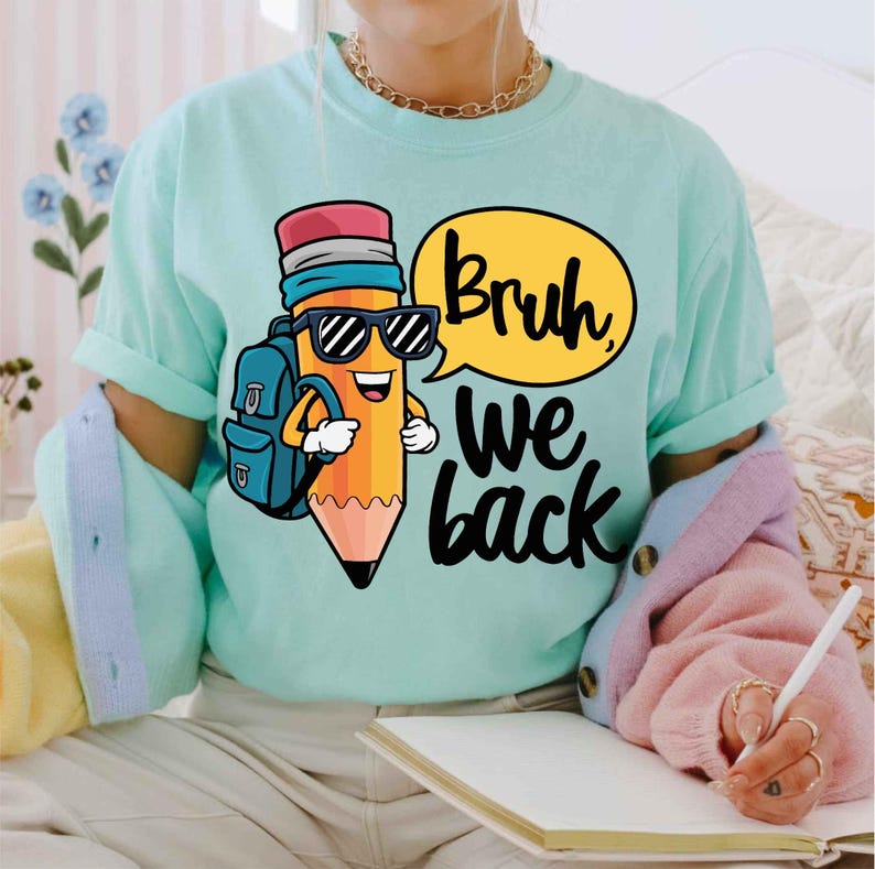 May include: A light teal t-shirt featuring a cartoon pencil character wearing sunglasses and a backpack. The pencil has a speech bubble that says "Bruh, we back" in black text. The shirt is a comfortable, casual style.