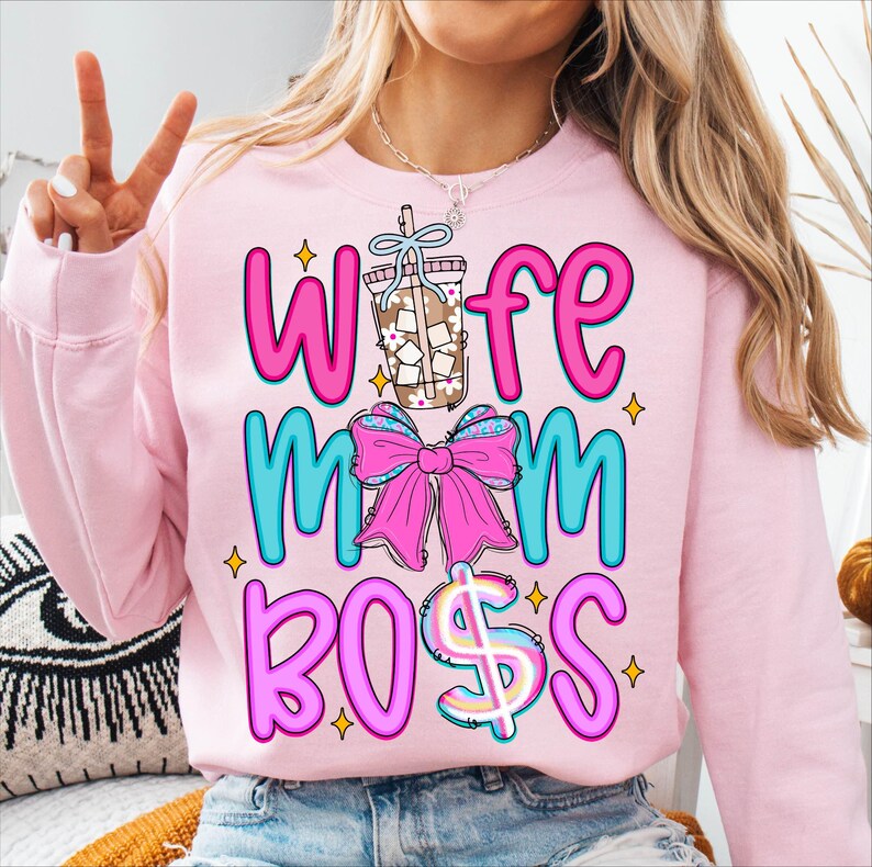 May include: Pink sweatshirt with the words "Wife Mom Boss" in a colorful design. The word "Wife" is in pink, "Mom" is in blue, and "Boss" has a dollar sign. A graphic of a drink and a pink bow are also featured.