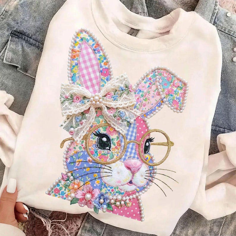 May include: Cream-colored sweatshirt featuring a colorful bunny design. The bunny has floral patterned ears, a bow, and round glasses. The design incorporates pink, blue, and yellow floral patterns, with pearl accents.