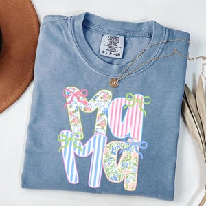 May include: A blue Comfort Colors t-shirt with the word "Mama" in a floral and striped design. The letters are decorated with bows in pink, green, and blue. A gold necklace with a pendant is draped across the shirt.