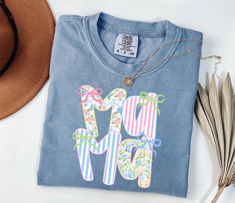 May include: A blue Comfort Colors t-shirt with the word "Mama" in a floral and striped design. The letters are decorated with bows in pink, green, and blue. A gold necklace with a pendant is draped across the shirt.