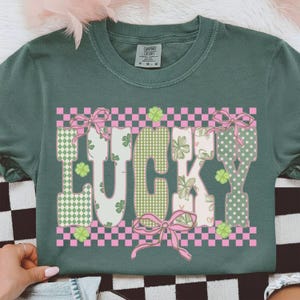May include: A dark green t-shirt with the word "LUCKY" in a decorative font. The letters are patterned with green gingham, polka dots, and floral designs, accented with pink bows and shamrocks. The shirt has a crew neck and short sleeves.