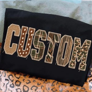 Op de afbeelding: Een zwart t-shirt met het woord CUSTOM in grote applicatieletters. Elke letter is gemaakt van een andere stof, waaronder beige jute, bruine stippen, groen ruitpatroon, camouflage, houtnerf en een bosrijk bladmotief.