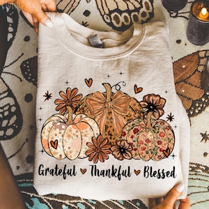 May include: A beige sweatshirt featuring a fall-themed design. The design includes three pumpkins with floral and leopard print patterns, surrounded by flowers and hearts. The words "Grateful, Thankful, Blessed" are printed below the pumpkins.