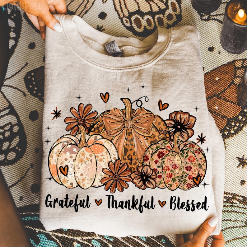 May include: A beige sweatshirt featuring a fall-themed design. The design includes three pumpkins with floral and leopard print patterns, surrounded by flowers and hearts. The words "Grateful, Thankful, Blessed" are printed below the pumpkins.