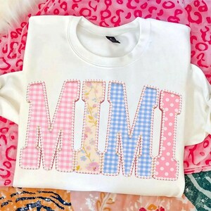 May include: White sweatshirt with the word "MIMI" in large, colorful letters. The letters are decorated with pink gingham, floral patterns, blue gingham, and pink polka dots, outlined with small white beads.
