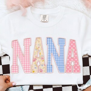 May include: White crew neck sweatshirt with the word "NANA" in large, colorful letters. The letters are patterned with pink gingham, floral, blue gingham, and pink polka dots, outlined with small pearls.