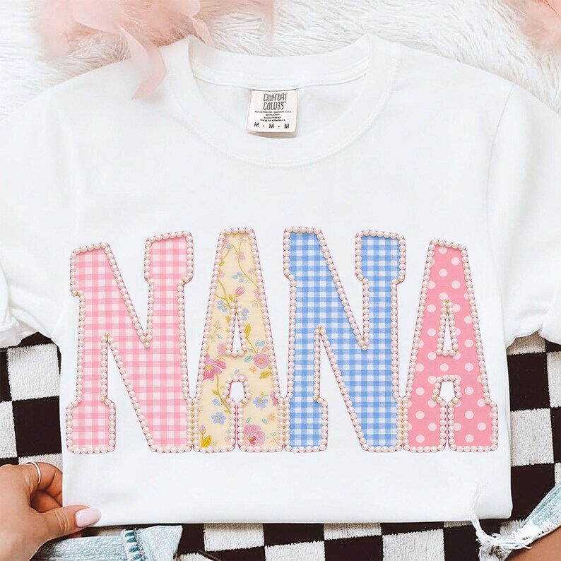 May include: White crew neck sweatshirt with the word "NANA" in large, colorful letters. The letters are patterned with pink gingham, floral, blue gingham, and pink polka dots, outlined with small pearls.