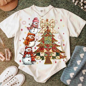 May include: White t-shirt featuring a Christmas tree design with a large bow and three snowmen wearing hats and scarves. The tree is decorated with red, green, and gold patterns. Red star accents are scattered around the tree and snowmen.