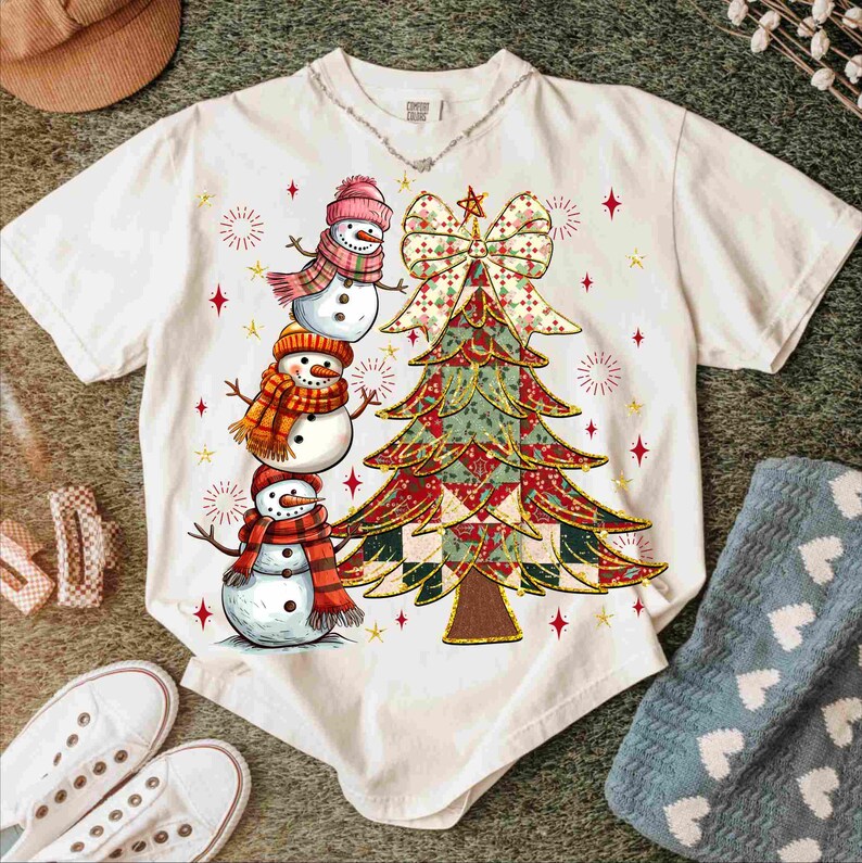 May include: White t-shirt featuring a Christmas tree design with a large bow and three snowmen wearing hats and scarves. The tree is decorated with red, green, and gold patterns. Red star accents are scattered around the tree and snowmen.