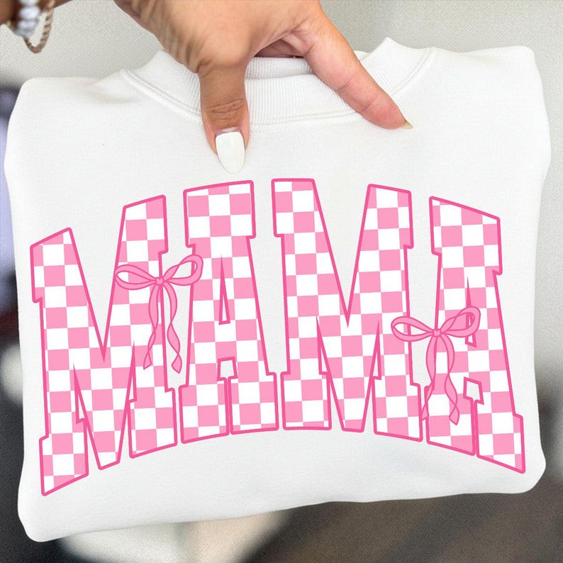 May include: White sweatshirt with the word "MAMA" in large, pink, checkered letters. The letters have a collegiate style font and are accented with pink bows. The sweatshirt is folded and held up by a hand.