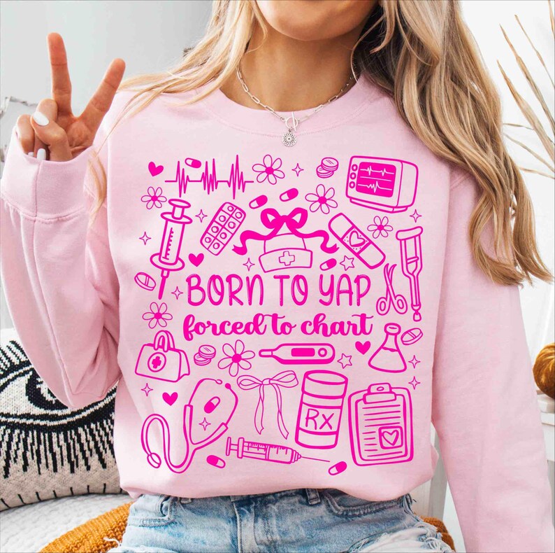 May include: Pale pink sweatshirt with a pink graphic design featuring medical symbols, flowers, and the text "BORN TO YAP forced to chart". The design includes a stethoscope, pills, and a nurse's hat.
